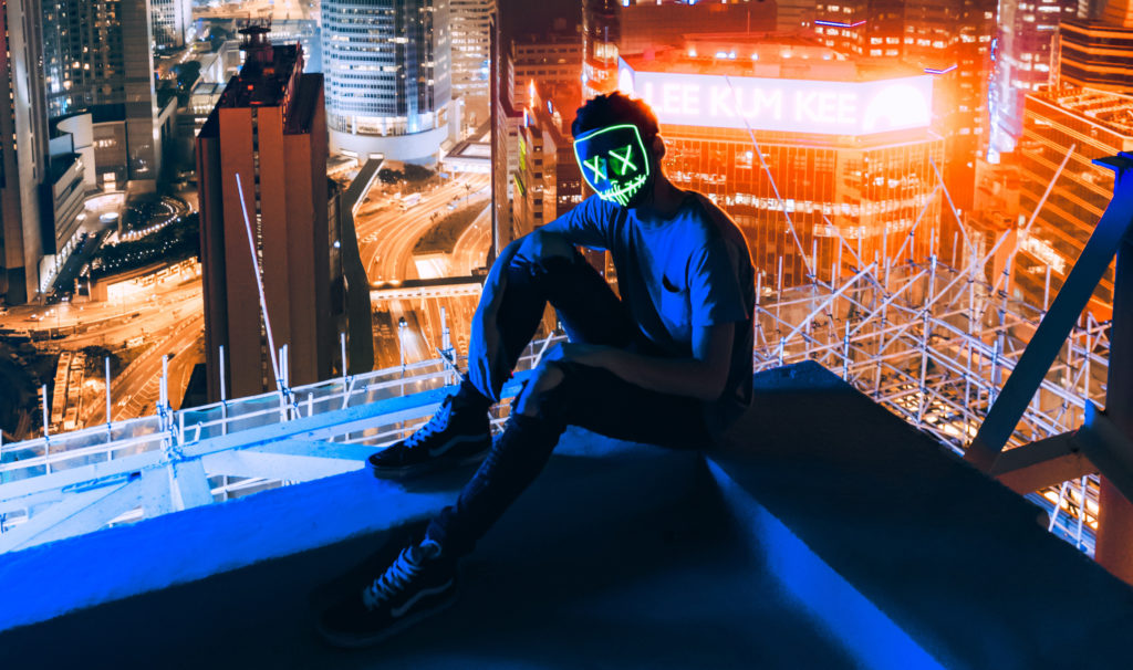 A human sits in shadow atop a skyscraper's rooftop overlooking a brightly lit neon cityscape. They wear a mask designed with LED lights showing Xs for eyes and a skeletal smile.