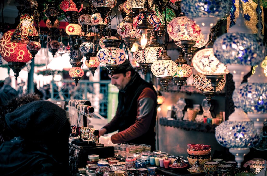 The interior of a shop, a human male at the counter, colorful lamps hanging from the ceiling and the counter stuffed full of colorful ceramics and bands.