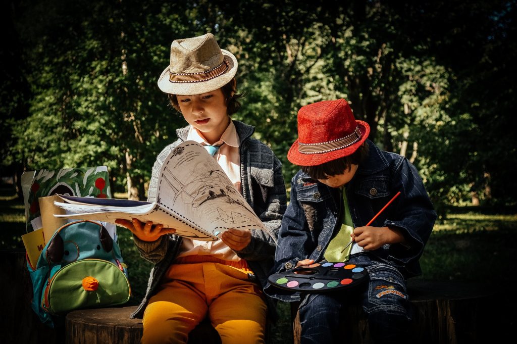 Two young boys painting and sketching with reference manuals while sitting on tree stumps in a lush, green park.