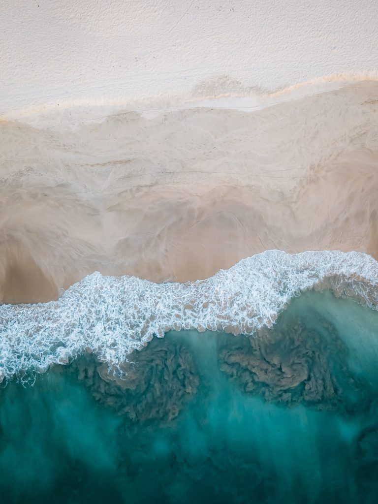 A foam of white ocean water cresting a wave overtakes a bright beach, carrying clear blue water up onto the shore.