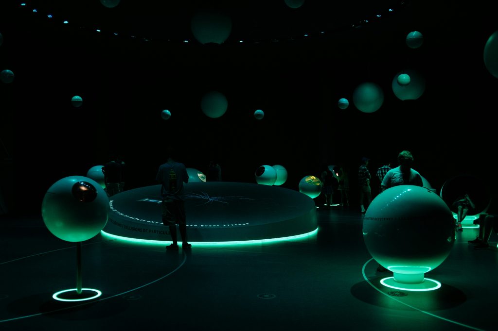 Humanoid figures explore a dark room, filled with futuristic orblike consoles and a central station lit with soft green hues.