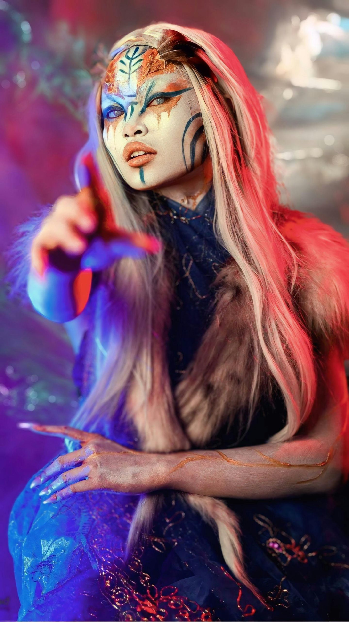 An alien looking figure with intricate facepaint points toward the viewer. They're lit with blue and red hues.