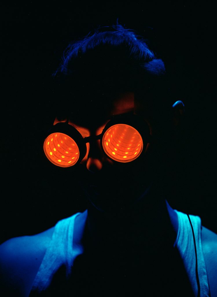 The ominous silhouette of a humanoid figure is cast in shadow. The only source of light or color comes from a pair of orange-glowing science goggles that obscure the wearer's facial features and a soft blue glow across their shoulders.