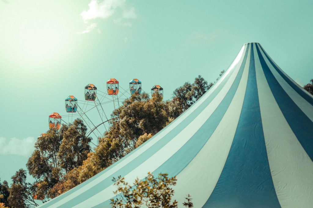Ferris wheel partially visible against a pale blue sky beside a large blue-and-white striped circus tent.