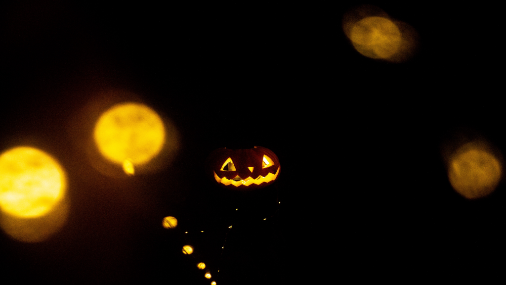 Over a dark background, yellow orb lights float around an illuminated jack-o-lantern in the center of the screen.