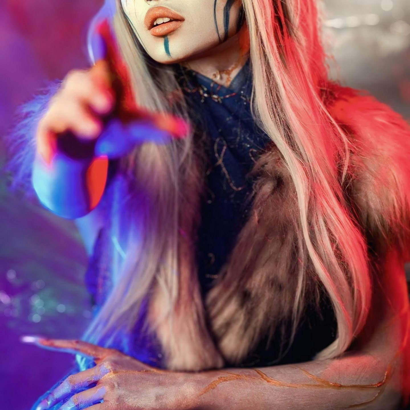 An alien looking figure with intricate facepaint points toward the viewer. They're lit with blue and red hues.